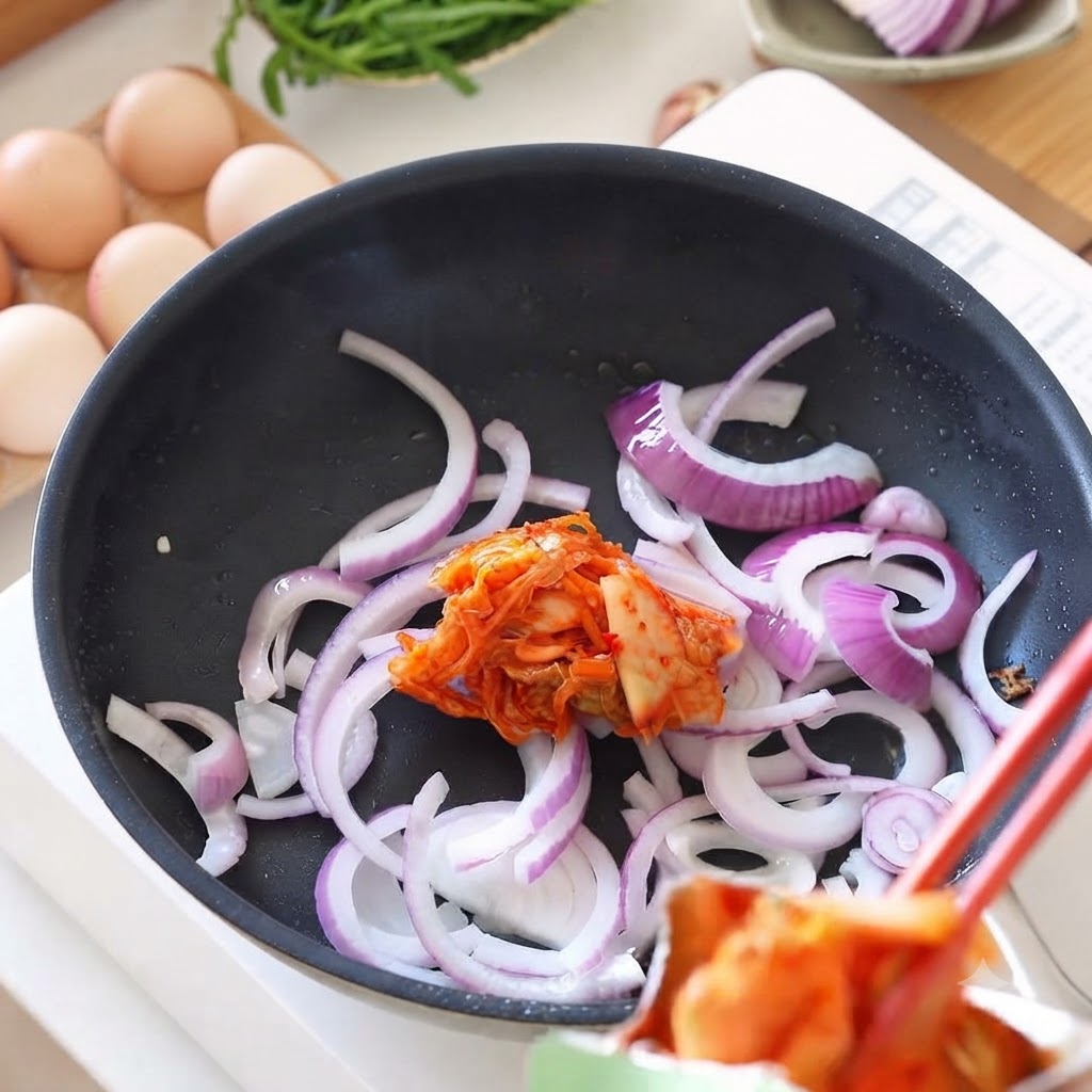 Frisches Kimchi wird mit Stäbchen in eine Pfanne mit angeschmorten Zwiebeln gegeben.