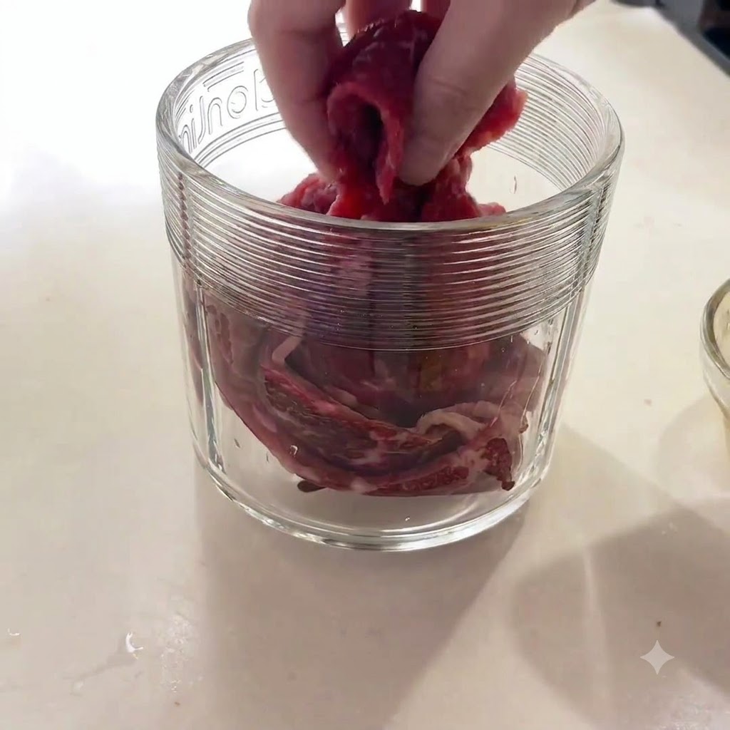 Eine Hand legt rohe Rindfleischstücke in den Glasbehälter einer Küchenmaschine.
