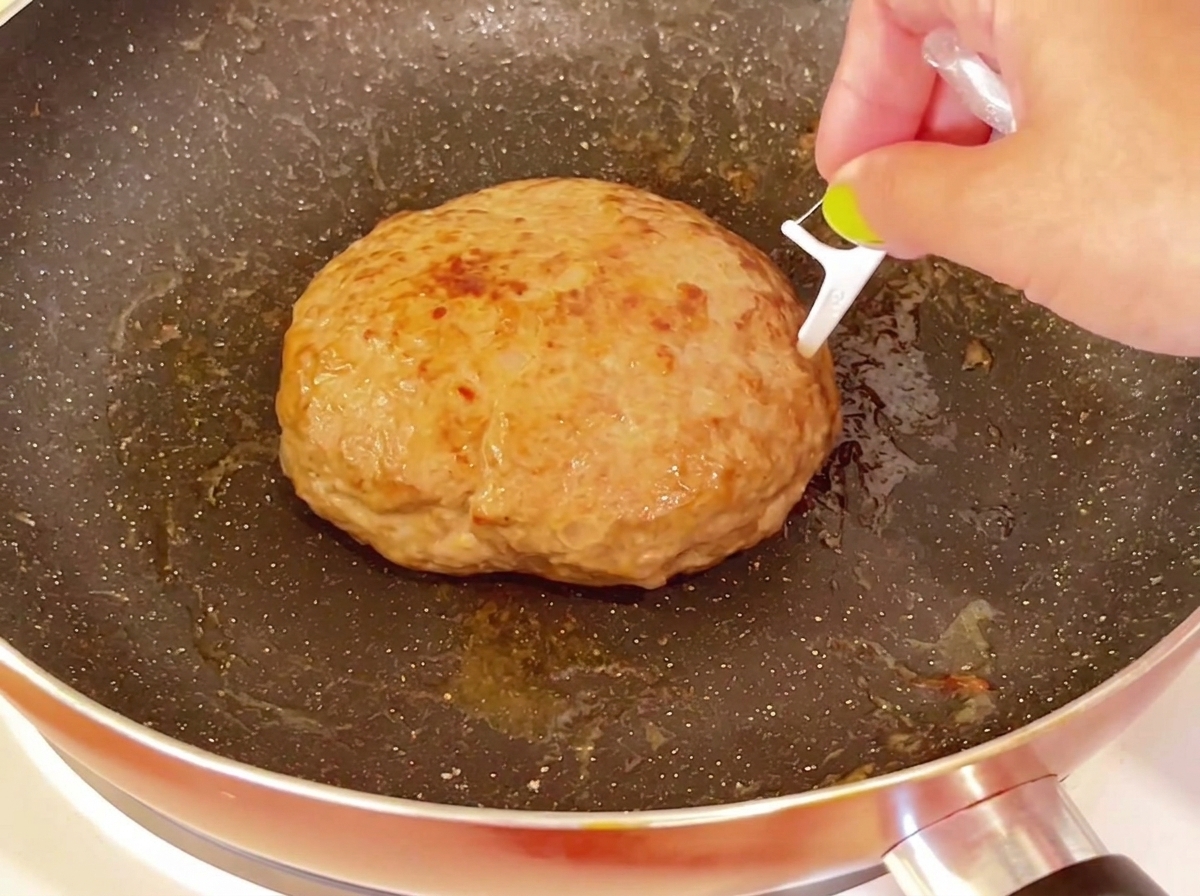 Ein Zahnstocher wird in die obere Fläche eines dicken, gebratenen Hamburger-Pattys in einer Pfanne gestochen.