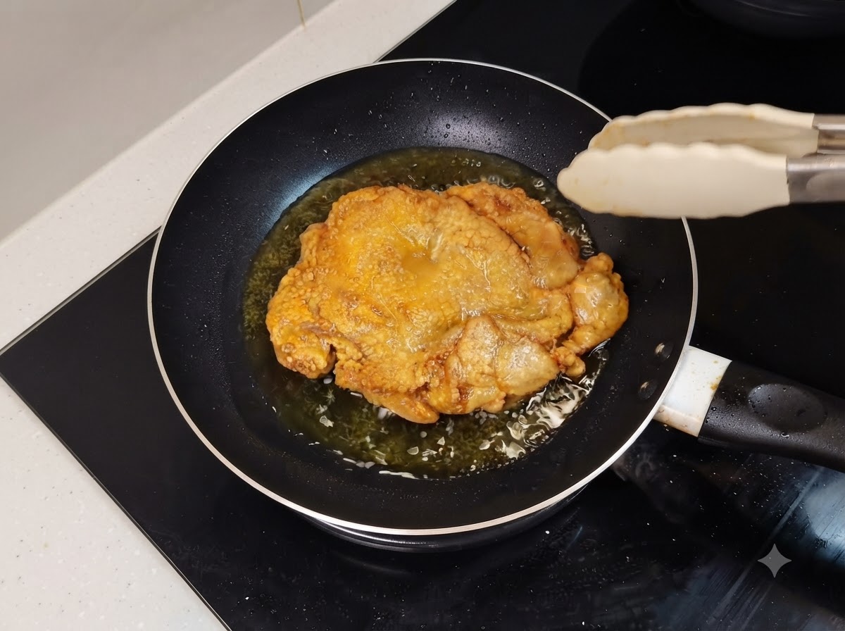Ein goldbraun gebratener Hähnchenschenkel, der in einer öligen Bratpfanne zischt, mit Küchenzange in der Nähe.