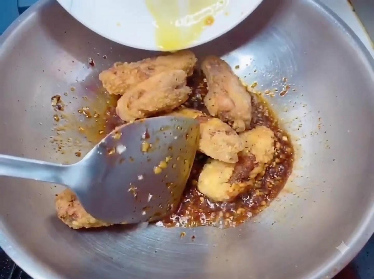 Knusprig goldene frittierte Hähnchenflügel werden mit einem Spatel in den Wok mit der Soße gegeben.