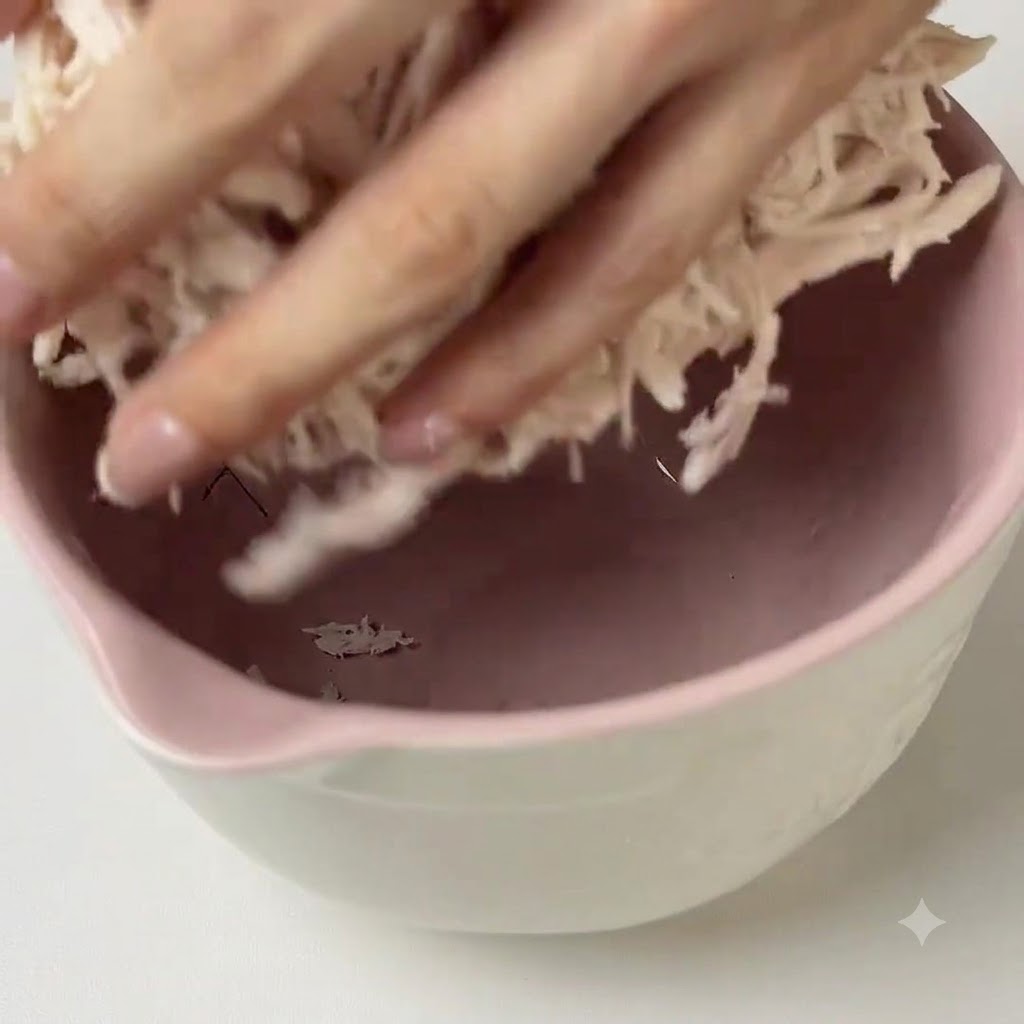 Hände vermengen grünes Pesto mit gezupftem Hähnchen in einer rosa Rührschüssel.