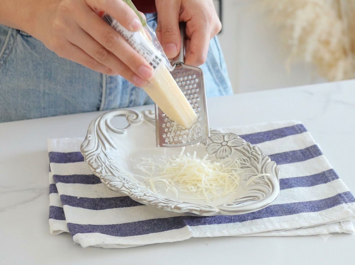 Parmesankäse, der frisch mit einer Metallreibe auf einen weißen Teller gerieben wird.