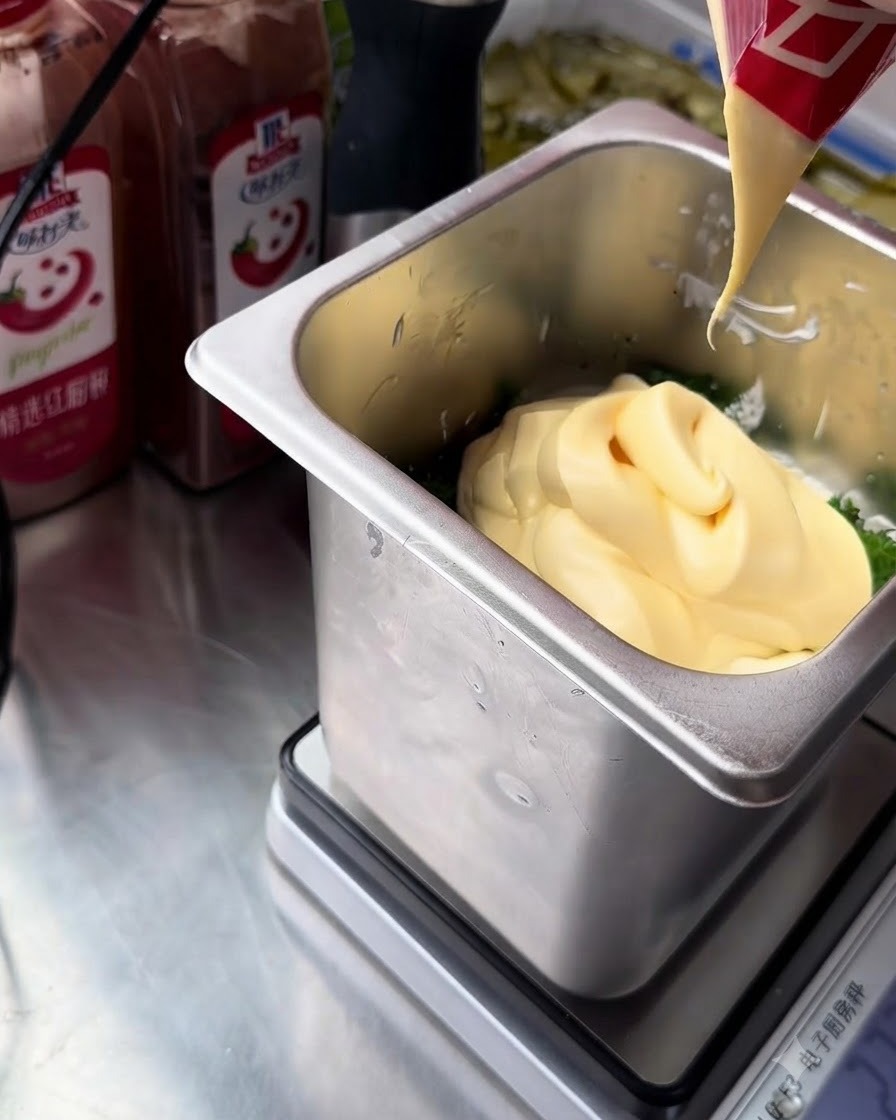Ein Strahl reichhaltiger, gelber Eigelb-Mayonnaise wird aus einer rot-weißen Verpackung in den Edelstahlbehälter mit den Tartarsaucenzutaten gedrückt.